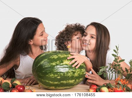 Three children sitting at the table. The boy put his hand on a large watermelon and whispers in his ear a girl. Gray background. Close-up.