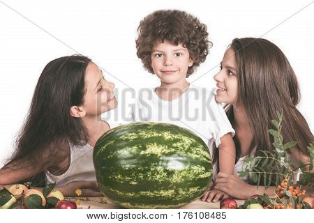 Three children sitting at the table in front of a large watermelon whole. The boy looks at the camera. The girls look at each other. White background. Close-up.