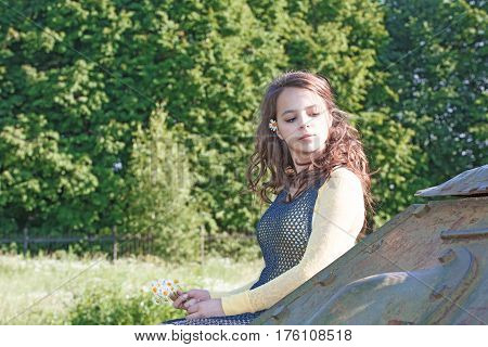 Girl with her hair sitting on a tank armor. In the hands holding a daisy flowers. Against the background of green forest. Close-up.