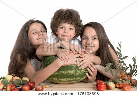 Brother and two sisters embrace a large watermelon sitting at the table. Laughing and looking into the camera. White background. Close-up.