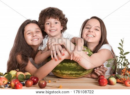 Brother and two sisters hugging a huge watermelon sitting at a table laughing looking at the camera. White background. Close-up.
