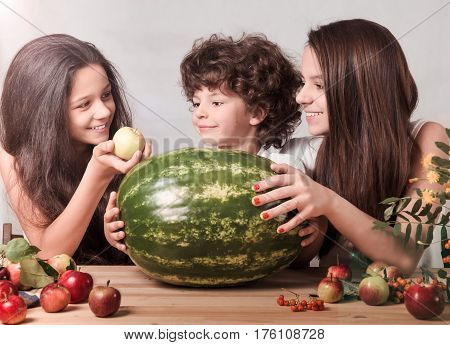 Three children playing with fruits sitting at the table in front of a huge watermelon. Gray background. Close-up.