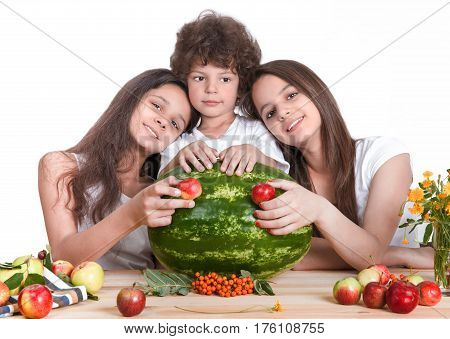 Three happy children are playing with a huge watermelon and fruit sit at the table. The sisters put their heads on the shoulders of his brother. White background. Close-up.