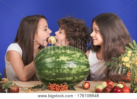 Two sisters and little brother sitting at the table. On the table is a huge whole watermelon and fruit. Brother and sister biting one apple. Close-up.
