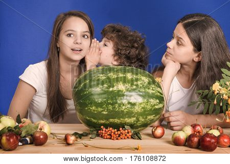 Kids talking while sitting at the table in front of a large watermelon whole. Looking at the camera. Blue background. Close-up.