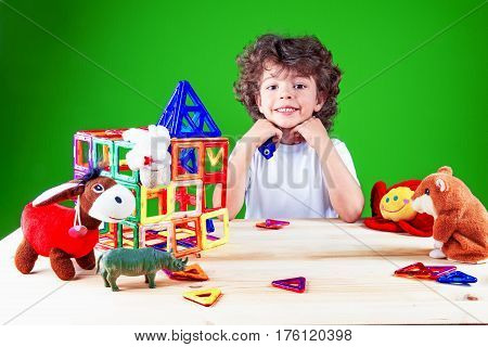 Widely smiling brown-eyed boy close up in white t-shirt looking at the camera. Near built home surrounded by toys.The construction of the house. Toys help to build the house. On a green background.