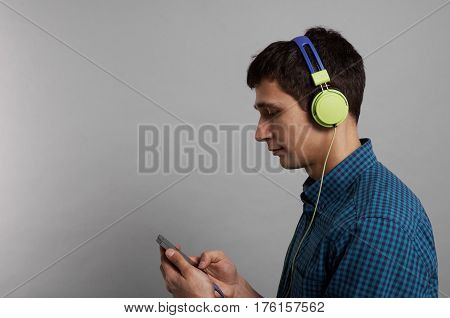 Side shot of student instudio with grey background. Man keep phone use internet technology and listen music in green headphones. Copy space for a text or advertising products.
