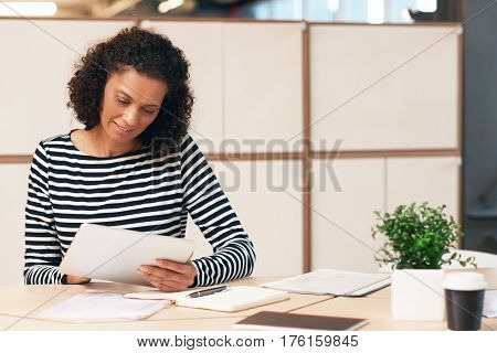 Confident and focused businesswoman wearing casual clothes sitting at a desk in a modern office organizing paperwork and working on a digital tablet