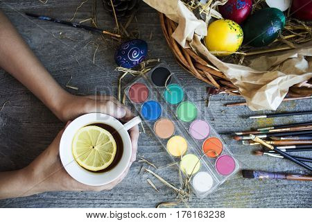 Childs Hand Holdind Cap Of Tea With Lemon And Next To Him Paints And Brush For Coloring Eggs For Eas