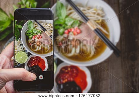 Taking a photo by Finger Pressing on Smartphone for Photograph Pho Vietnamse Noodle Soup on Wooden Background with Copy Space. Image for Food Advertise Concept