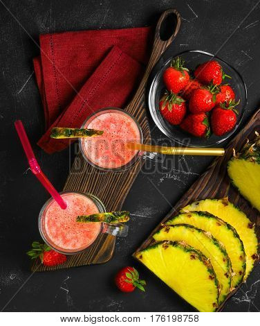 Summer Fruit cocktail smoothie with strawberries and pineapple in the glasses. Ingredients for cocktail smoothie of fresh strawberries and pineapple. Dark black background. Top view from above and copy space.