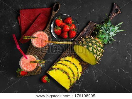 Summer Fruit cocktail smoothie with strawberries and pineapple in the glasses. Ingredients for cocktail smoothie of fresh strawberries and pineapple. Dark black background. Top view from above and copy space.