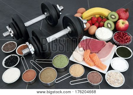 Health food for body builders with lean meat, salmon, dairy, fruit, nuts, pulses, seeds, supplement powders and dumbbell weights on slate background.