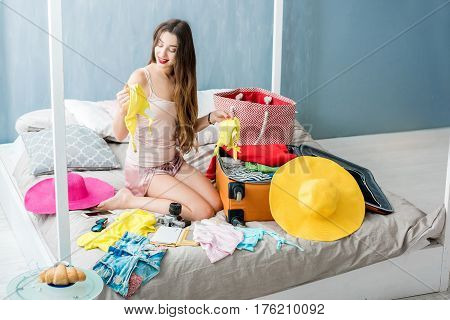 Young woman choosing swimsuit sitting with suitcase and clothes on the bed. Prepairing for the summer vacations