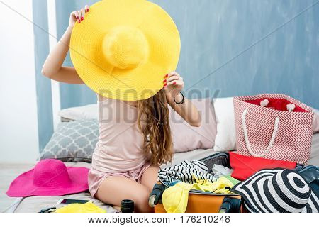 Young woman choosing hat sitting with clothes and suitcase on the bed. Prepairing for the summer vacations