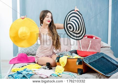 Young woman choosing hat sitting with clothes and suitcase on the bed. Prepairing for the summer vacations