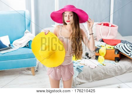 Young woman choosing hat in the bedroom with suitcase and clothes on the background. Prepairing for the summer vacations