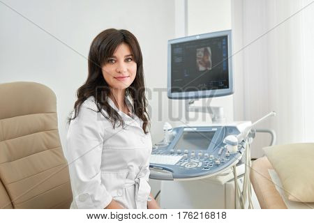 Professional in white uniform in modern clinic smiling at camera. Attractive brunette female doctor sitting in cabinet against ultrasound equipment with computer.