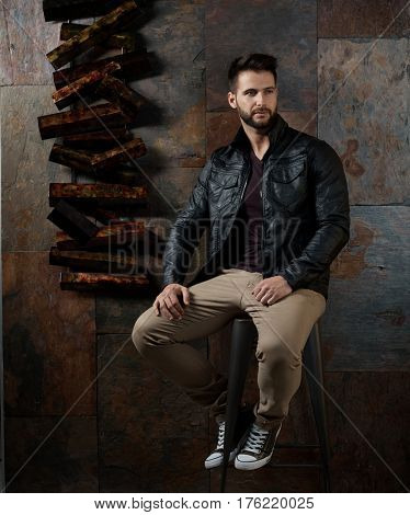 Man sitting on metal barstool against rusty wall, looking away.