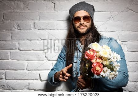 Portrait photo of rocker guy in sunglasses and woolly hat standing against brick wall holding bouquet of flowers for mother's day.