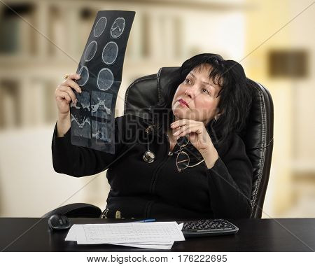 Female doctor takes brain x-ray image and scrupulous examines it. Black haired mature woman sits in tall office chair at black desk. Horizontal mid-shot on blurred indoors background