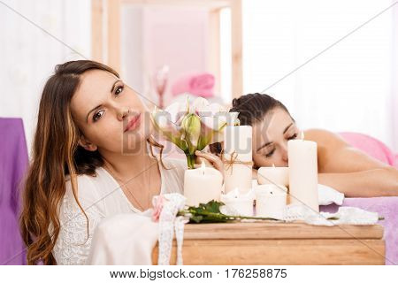 Messeur behind lighted candles, holding flowers with relaxing young woman on background at spa salon