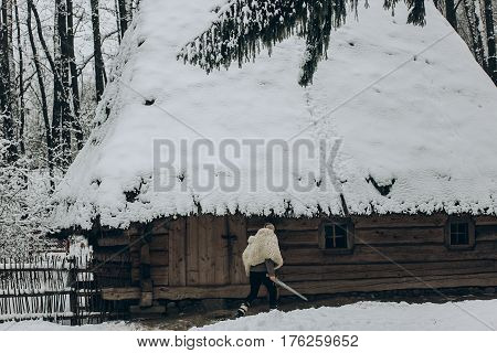 Strong Viking With Sword And Wolf Pelt Walking Towards Historical Wooden Building In North Woods In