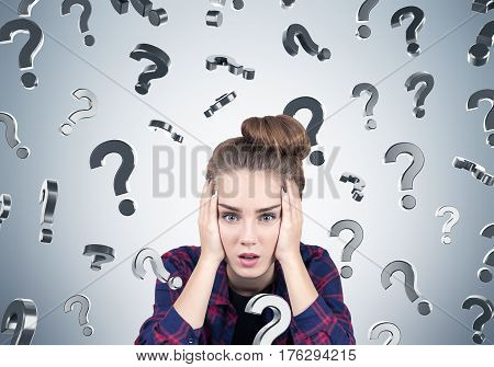 Portrait of a stressed teen girl sitting near a gray wall with question marks falling aground her. Concept of too many questions.