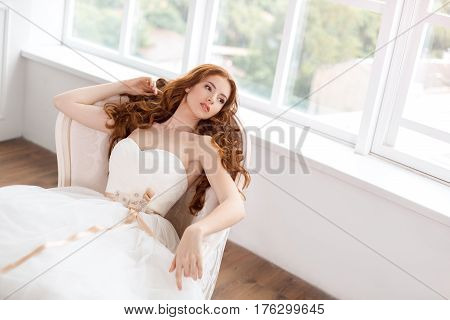 Bride in beautiful dress laying on sofa indoors in white studio interior like at home. Trendy wedding style shot in full length. Young attractive caucasian red hair model like a bride against big window tender resting.