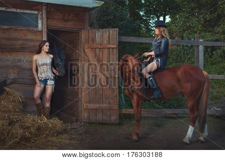 Woman came to the stables on horseback another woman takes the horse out of the stables.