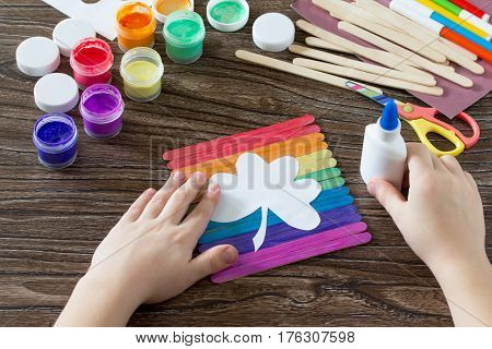 The Child Glues The Detail. Present For St. Patrick's Day Clover On A Rainbow, A Craft Of Of Wooden 