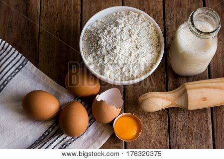 Eggs cracked shell open yolk flour milk ingredients for baking rolling pin on wood kitchen table linen towel top view