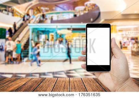 Mock up image of hand holding black mobile phone with blank white screen Empty wood table top on blurred images shopping mall for background perspective wood can be used for display products.