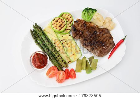 Grilled beef grilled with asparagus, zucchini, lobi, broccoli, tomato, chili and red sauce on a white plate, over white background