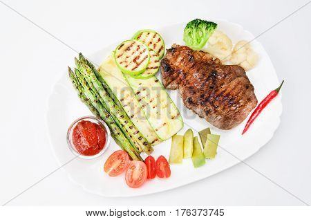 Grilled beef grilled with asparagus, zucchini, lobi, broccoli, tomato, chili and red sauce on a white plate, over white background