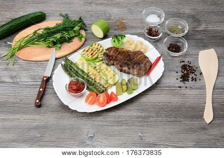 Grilled beef grilled with asparagus, zucchini, lobi, broccoli, tomato, chili and red sauce on a white plate, and other vegetables in wood background
