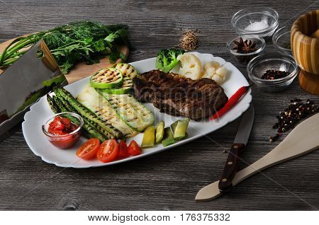 Grilled beef grilled with asparagus, zucchini, lobi, broccoli, tomato, chili and red sauce on a white plate, and other vegetables in wood background