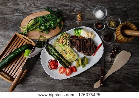 Grilled beef grilled with asparagus, zucchini, lobi, broccoli, tomato, chili and red sauce on a white plate, and other vegetables in wood background