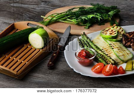 Grilled beef grilled with asparagus, zucchini, lobi, broccoli, tomato, chili and red sauce on a white plate, and other vegetables in wood background