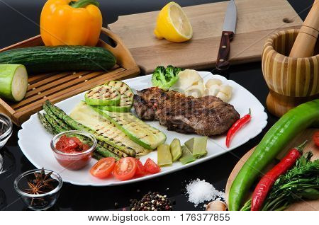 Grilled beef grilled steak with asparagus, zucchini, lobi, broccoli, tomato, chili and red sauce on a white plate, on a black glossy background
