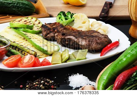 Grilled beef grilled steak with asparagus, zucchini, lobi, broccoli, tomato, chili and red sauce on a white plate, on a black glossy background