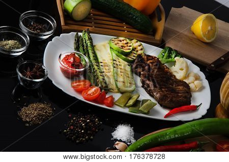 Grilled beef grilled steak with asparagus, zucchini, lobi, broccoli, tomato, chili and red sauce on a white plate, on a black glossy background