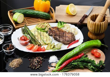 Grilled beef grilled steak with asparagus, zucchini, lobi, broccoli, tomato, chili and red sauce on a white plate, on a black glossy background