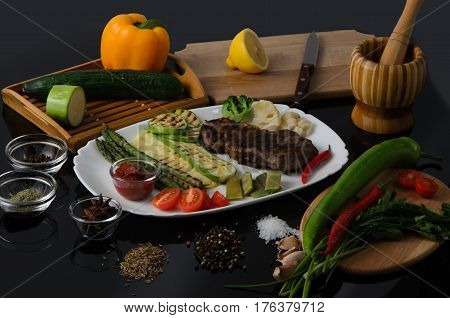Grilled beef grilled steak with asparagus, zucchini, lobi, broccoli, tomato, chili and red sauce on a white plate, on a black glossy background