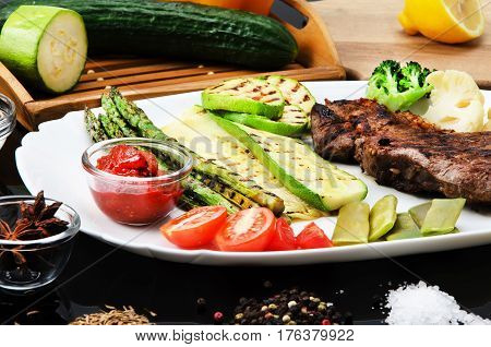 Grilled beef grilled steak with asparagus, zucchini, lobi, broccoli, tomato, chili and red sauce on a white plate, on a black glossy background