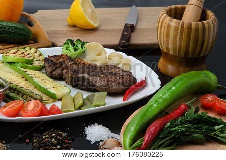 Grilled beef grilled steak with asparagus, zucchini, lobi, broccoli, tomato, chili and red sauce on a white plate, on a black glossy background