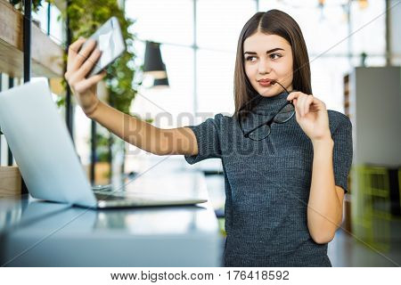 Young female taking a selfie in the office while sitting with laptop in modern office room