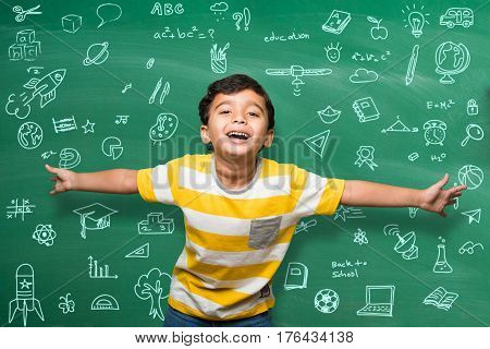 Indian school kid in hand stretched pose over green chalkboard or chalk board background, indian boy standing over green chalkboard background in the school with doodles -  back to school concept
