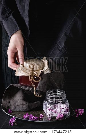 Femail hands hold glass jar of homemade lilac syrup. Glass jar of sugared lilac flowers and glass pitcher of lilac lemonade water on black tablecloth over black. Dark rustic atmosphere.