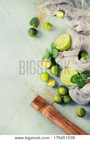Ingredients for mojito cocktail, whole, sliced lime and mini limes, mint leaves, brown crystal sugar over gray stone texture background with gauze textile and wooden bar muddler. Top view, space